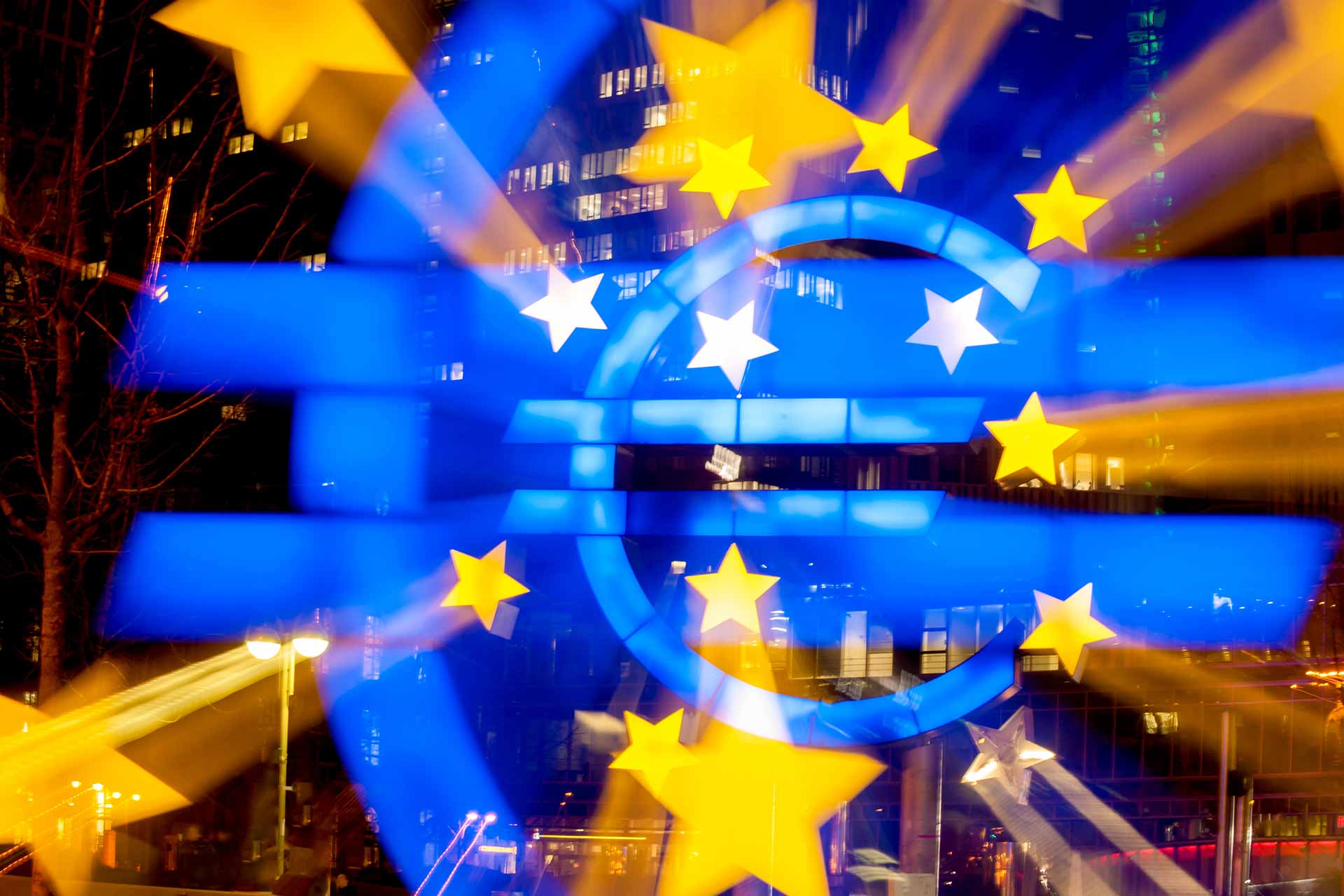 Leuchtendes Euro-Zeichen mit Sternen, im Hintergrund ein Finanzgebäude, EZB